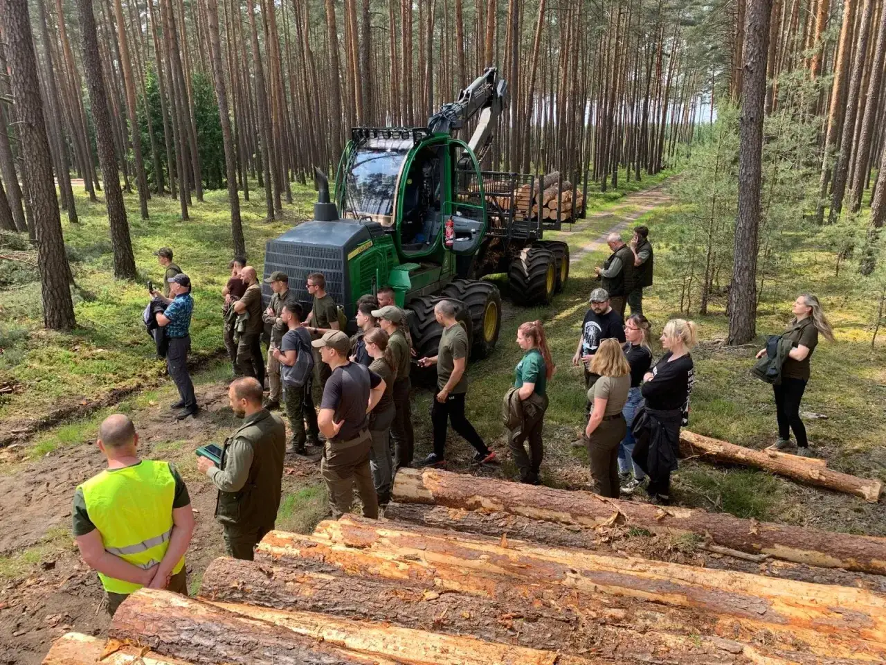 Grupa os&oacute;b na leśnej polanie, przyglądająca się maszynie leśnej. To część praktycznych zajęć w ramach studi&oacute;w leśniczych.