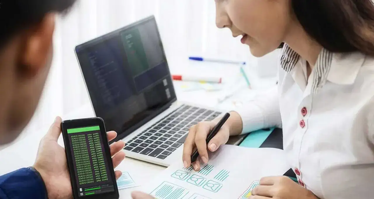 Tester aplikacji co to za praca? Dwoje ludzi analizuje kod na telefonie i laptopie, planując interfejs.