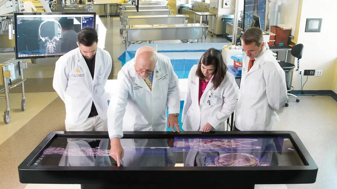 Profesor i studenci medycyny zaocznej analizują cyfrowy model anatomiczny na interaktywnym stole.