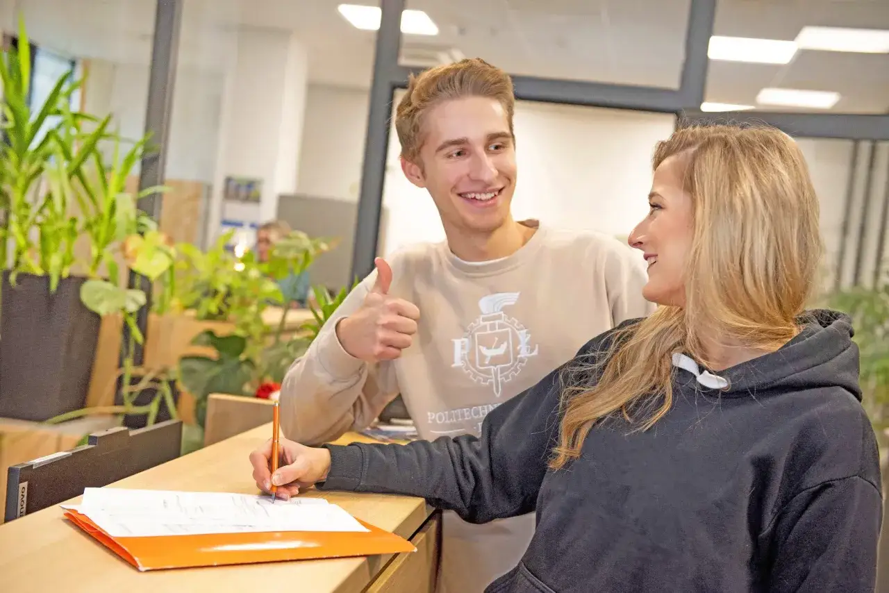 Uśmiechnięty student z kciukiem w górze wspiera koleżankę, która wypełnia dokumenty. To może być początek jej drogi na inną uczelnię.