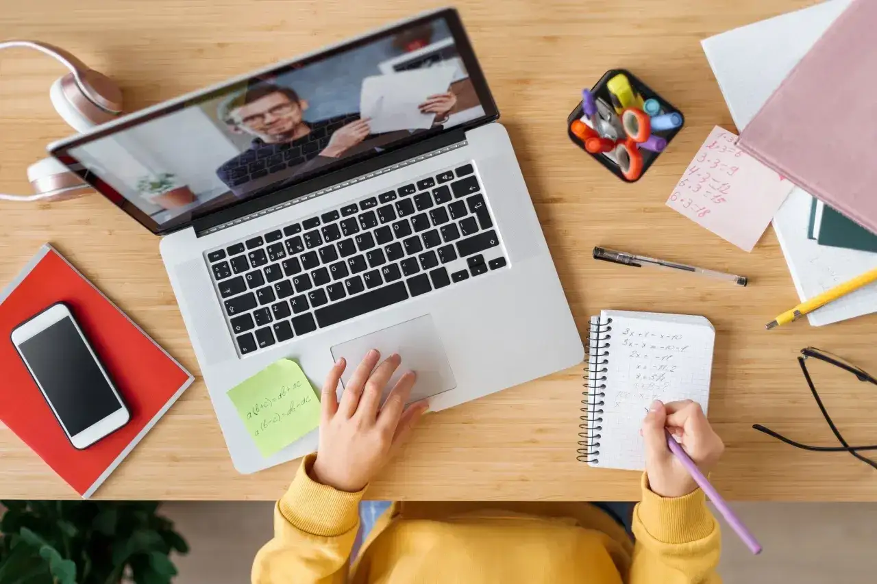 Uczeń uczy się zdalnie, korzystając z laptopa do studi&oacute;w hybrydowych. Na biurku leżą notatki, telefon i słuchawki.