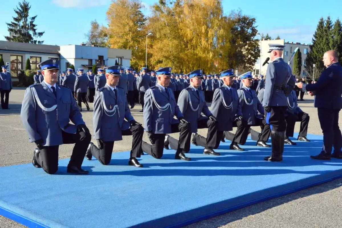 Uroczystość w szkole policyjnej. Policjanci w mundurach klęczą na niebieskim dywanie.